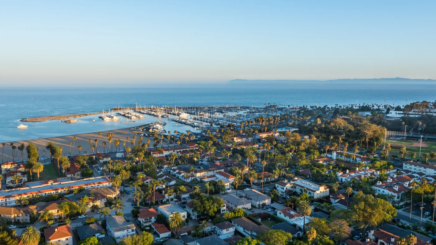 Aerial view over Santa Barbara at golden hour, looking across red-tile neighborhoods and the palm-lined beach to the harbor, with the Channel Islands on the horizon